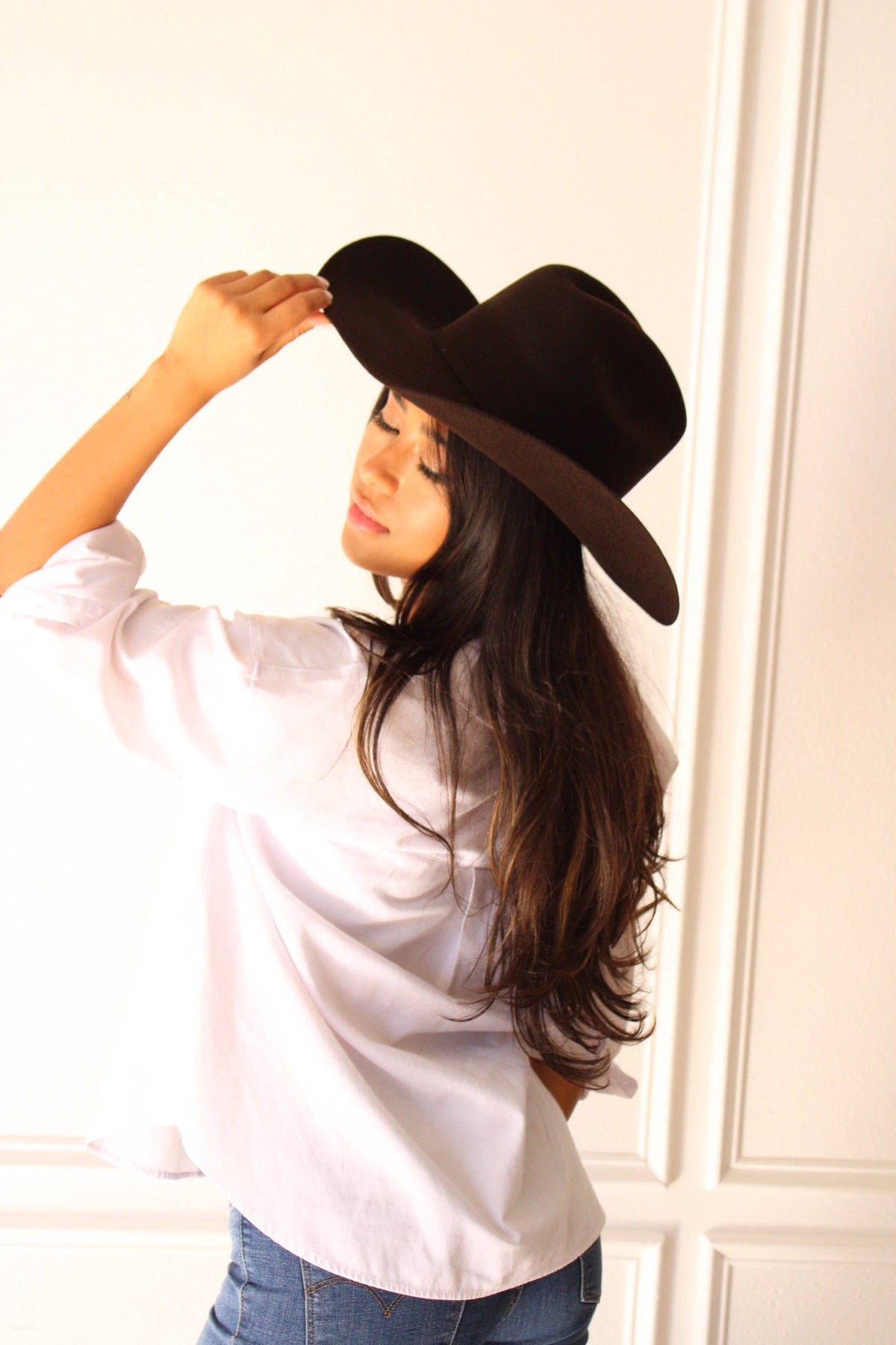 Model in a white shirt and jeans touching the brim of the chocolate brown Wren merino wool cowboy hat