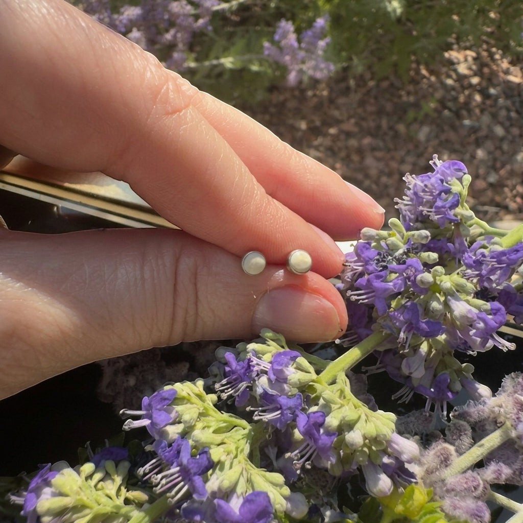Wildflower Artisans round mother of pearl stud earrings in sterling silver held by fingers near purple flowers