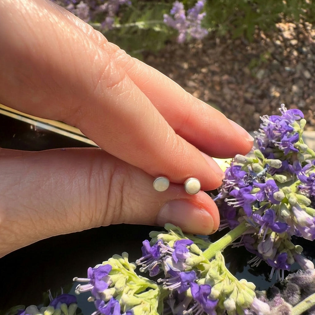 Wildflower Artisans round mother of pearl stud earrings in sterling silver held by fingers near purple flowers.