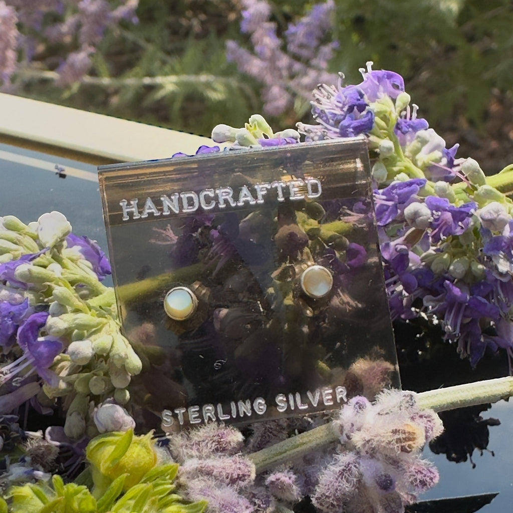 Wildflower Artisans round mother of pearl stud earrings in sterling silver settings displayed with purple flowers.