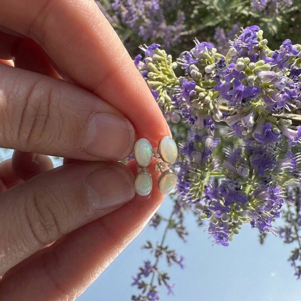 Wildflower Artisans oval opal stud earrings in sterling silver bezels held against a backdrop of purple flowers.