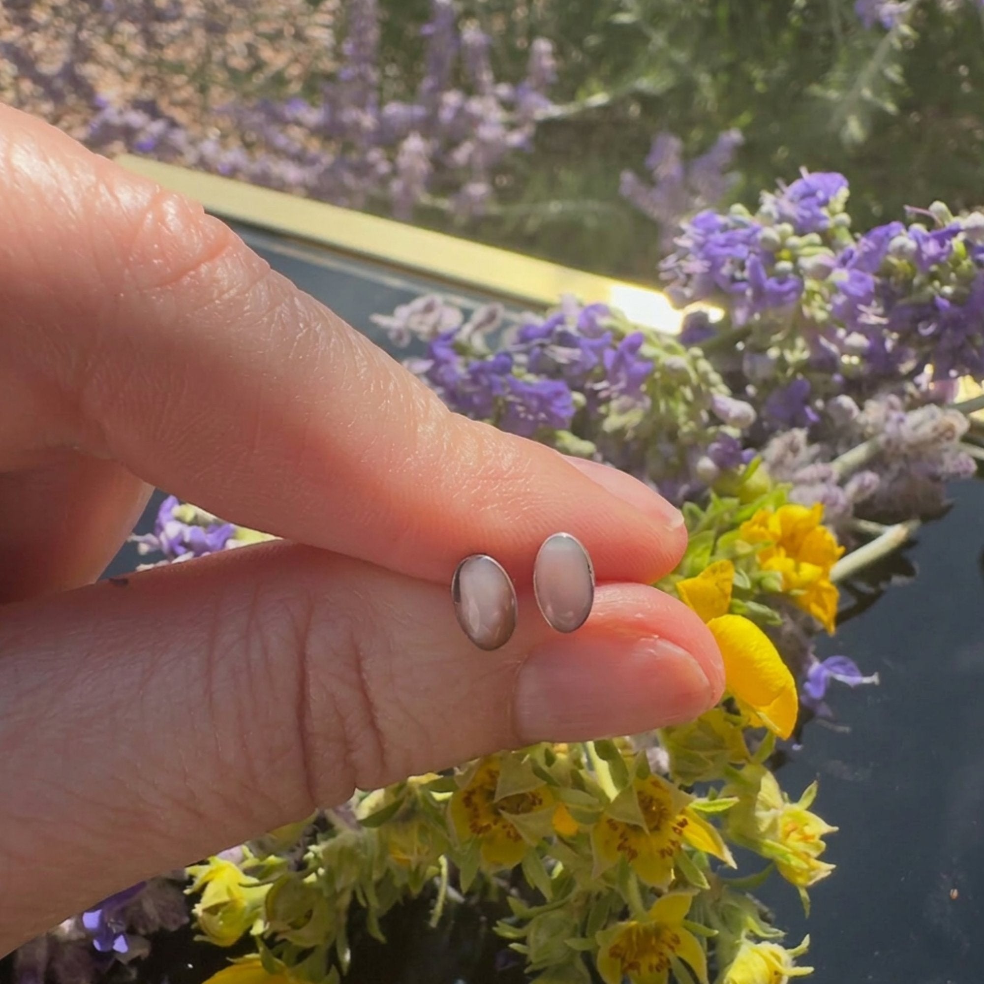 Wildflower Artisans oval opal stud earrings in sterling silver settings held between fingers against floral backdrop