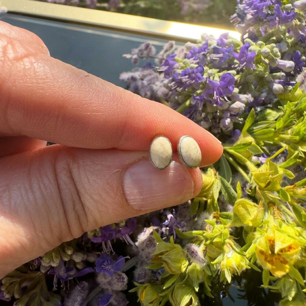 Wildflower Artisans oval stone stud earrings in sterling silver held by fingers against a backdrop of purple flowers