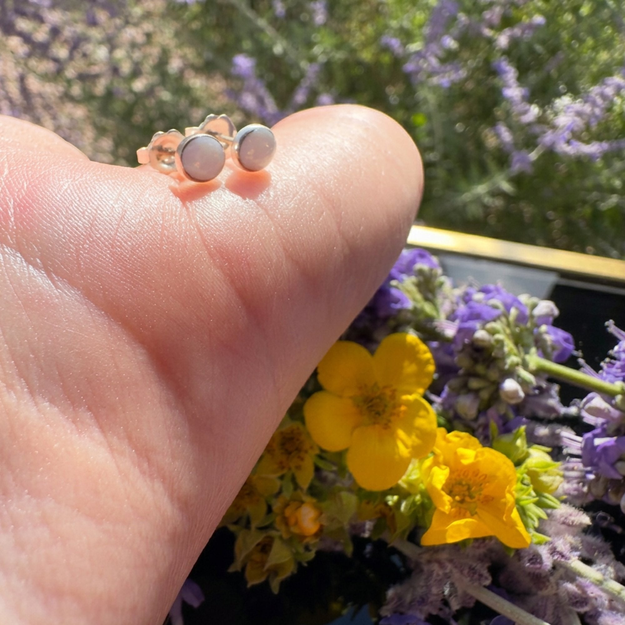 Wildflower Artisans round opal stud earrings set in sterling silver bezels held on a finger against floral backdrop