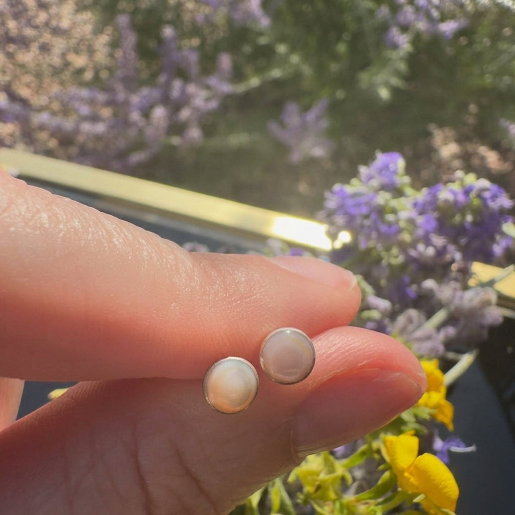 Wildflower Artisans round opal stud earrings in sterling silver bezels held between fingers against a floral backdrop.
