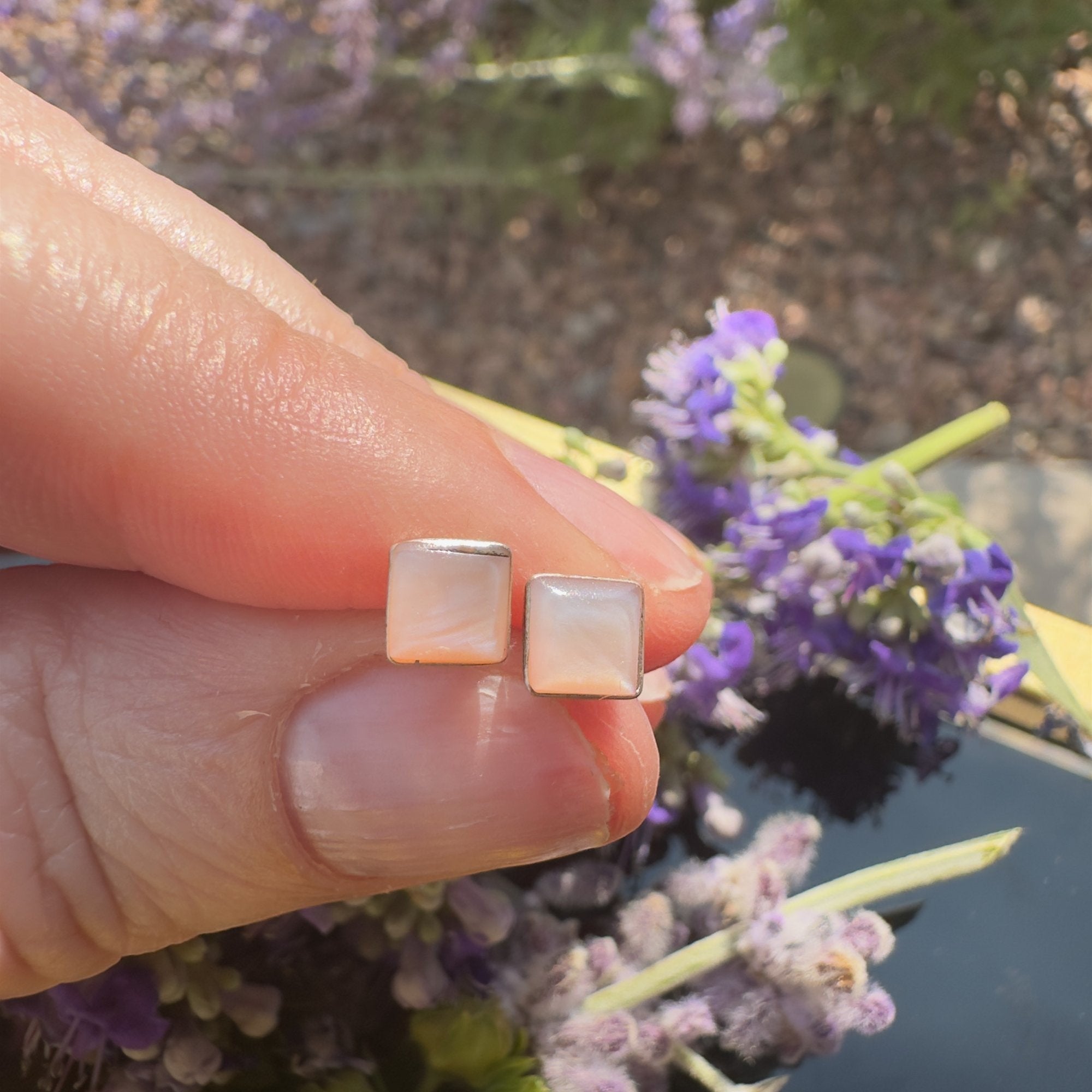 Wildflower Artisans square mother of pearl stud earrings in sterling silver settings held against purple flowers.