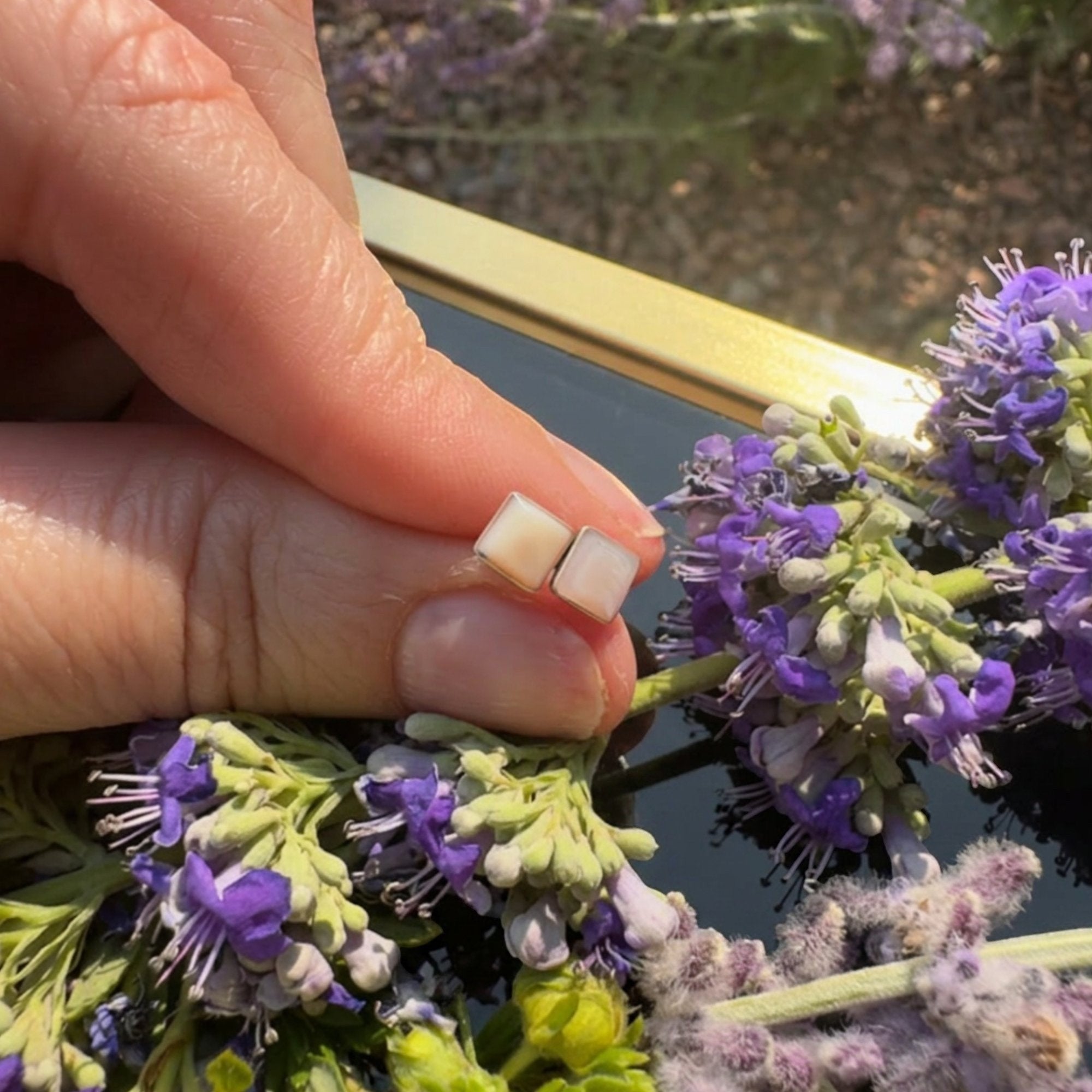 Wildflower Artisans square mother of pearl stud earrings in sterling silver held by hand near purple flowers